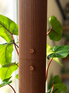 A close-up of a tall, ribbed brown cylindrical plant stand or tower, with two small hexagonal metal hooks on the front, surrounded by green houseplant leaves.